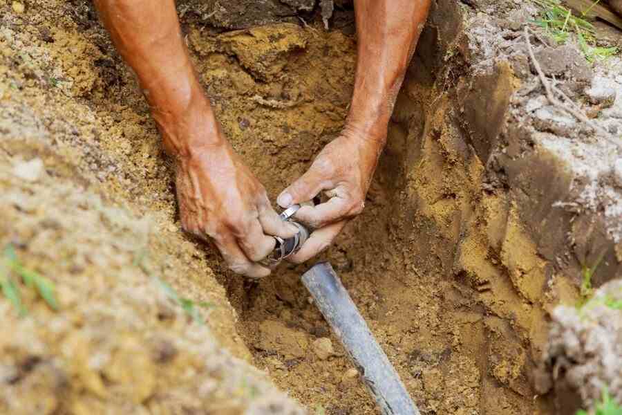 Worker's hands joining a fitting onto a black poly pipe at the bottom of a freshly dug trench.