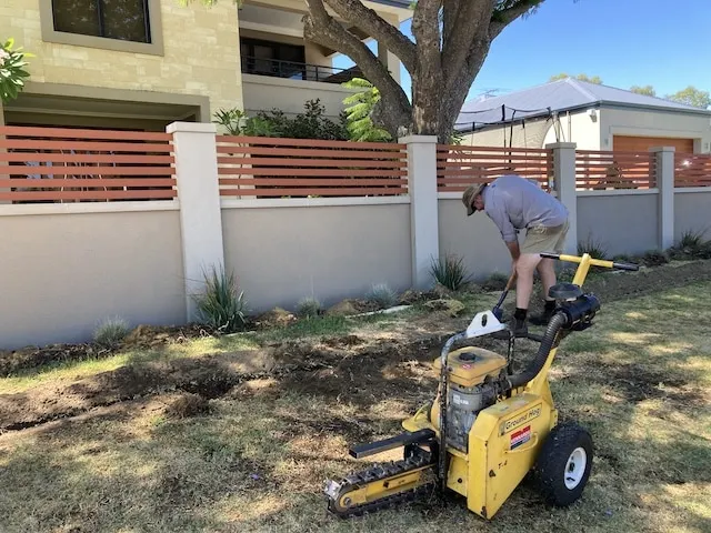 Tradesperson operating a yellow walk-behind trencher to cut a line across a front yard lawn.