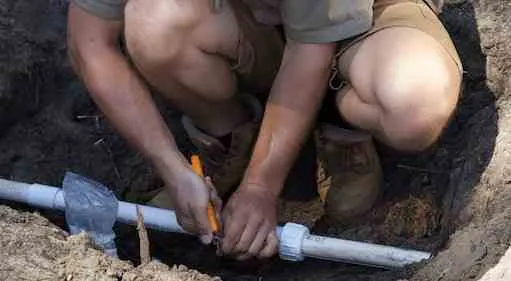 Worker crouched in a trench tightening a fitting onto a white PVC irrigation main with a wrench.