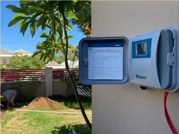 Hunter Hydrawise wifi controller mounted on an exterior wall beside its open instruction-covered enclosure.